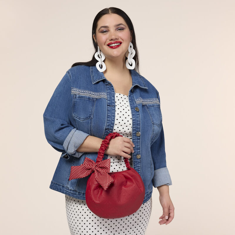 Woman smiling at camera wearing a white dress with black polka dots and denim jacket. She holds the Loungefly Minnie Mouse Rock the Dots Handbag over a wrist. 